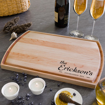 Light wood arched cutting board engraved with the Erickson family name and established date, styled on a table with champagne glasses and candles.