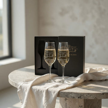 Two engraved champagne flutes with a pale sparkling drink beside a black gift box on a round stone table