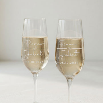 Two champagne flutes engraved with Romeo and Juliet and a date, filled with a pale sparkling drink against a light background.