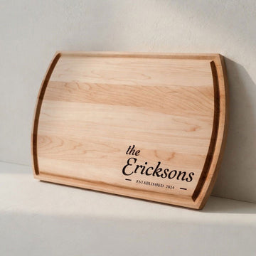 Maple cutting board engraved with the Ericksons and Established 2024, leaning against a light wall.