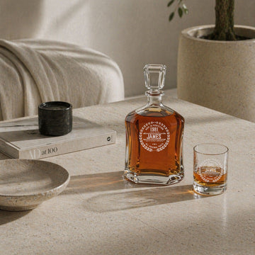 Personalized glass decanter and matching whiskey glass on a pale stone coffee table beside neutral seating.