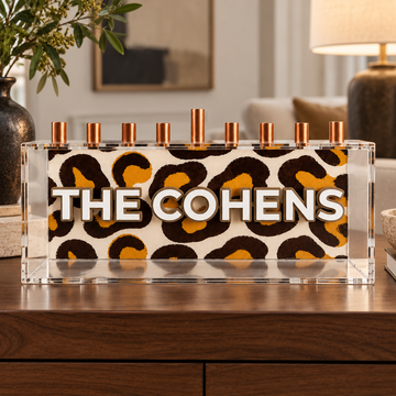 THE COHENS leopard-print family menorah with white text inside a clear acrylic box on a walnut console