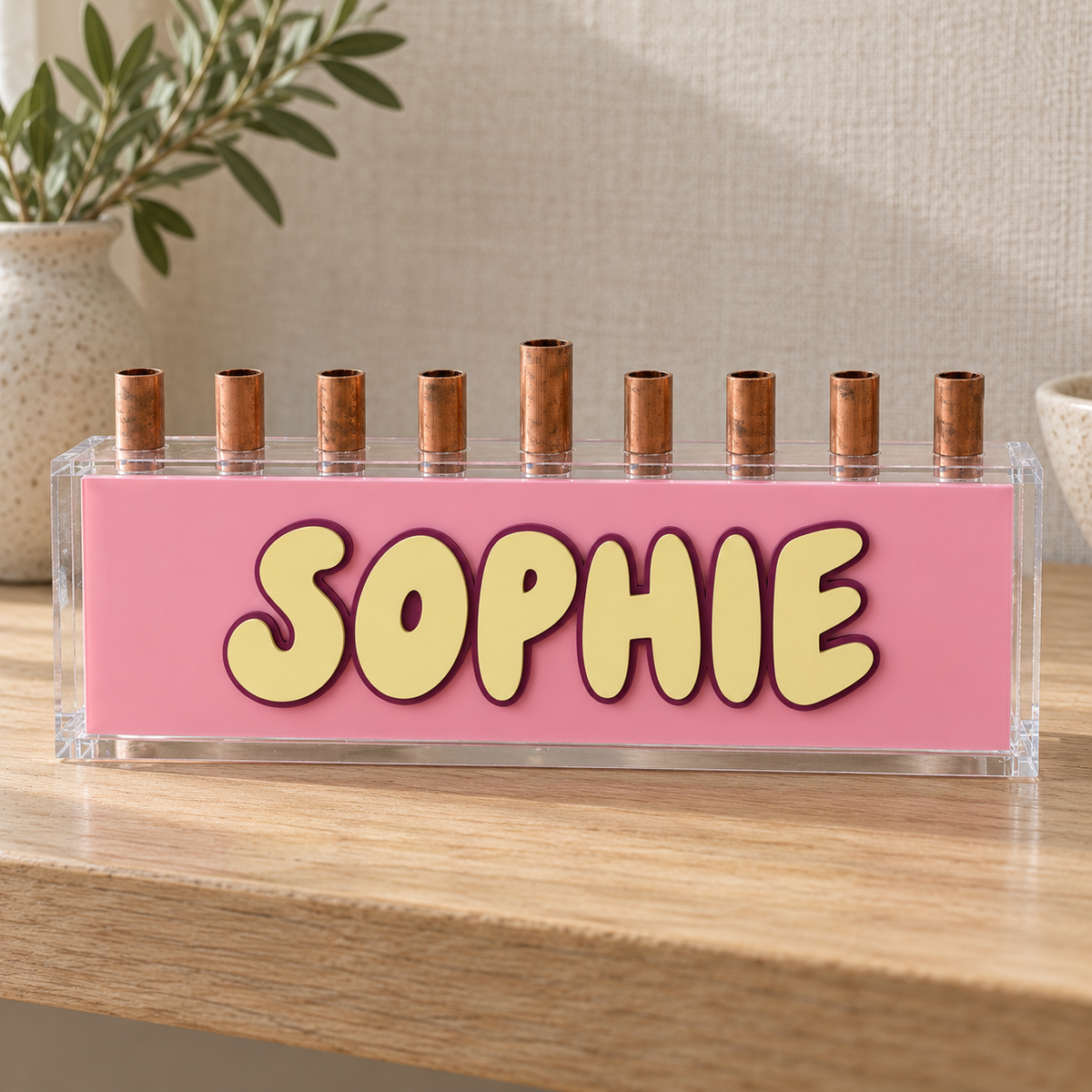 Pink Sorbet acrylic menorah with yellow SOPHIE lettering and raw copper candle tubes on an oak shelf
