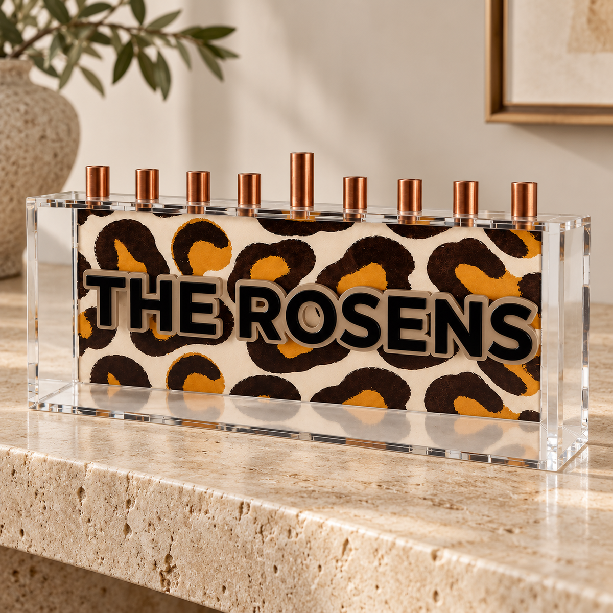 THE ROSENS leopard print menorah with black lettering inside a clear acrylic box on travertine